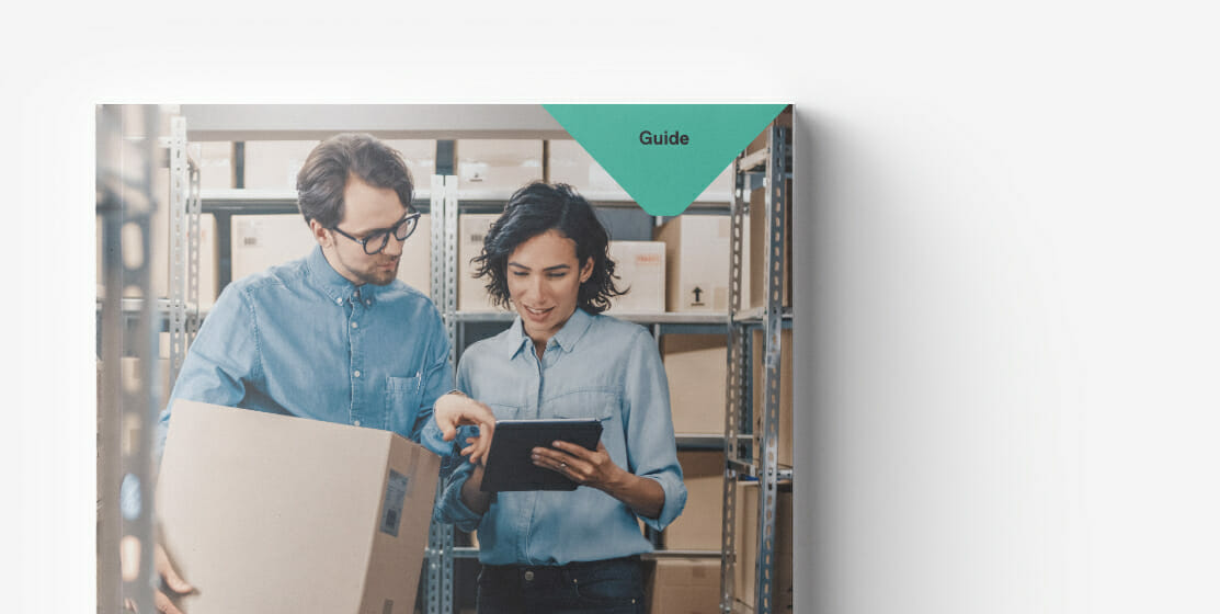 Two people stand in a storage area with shelves of boxes. One is holding a large box while the other points at a tablet, streamlining their B2B procurement process. A green triangular label in the corner says "Guide.