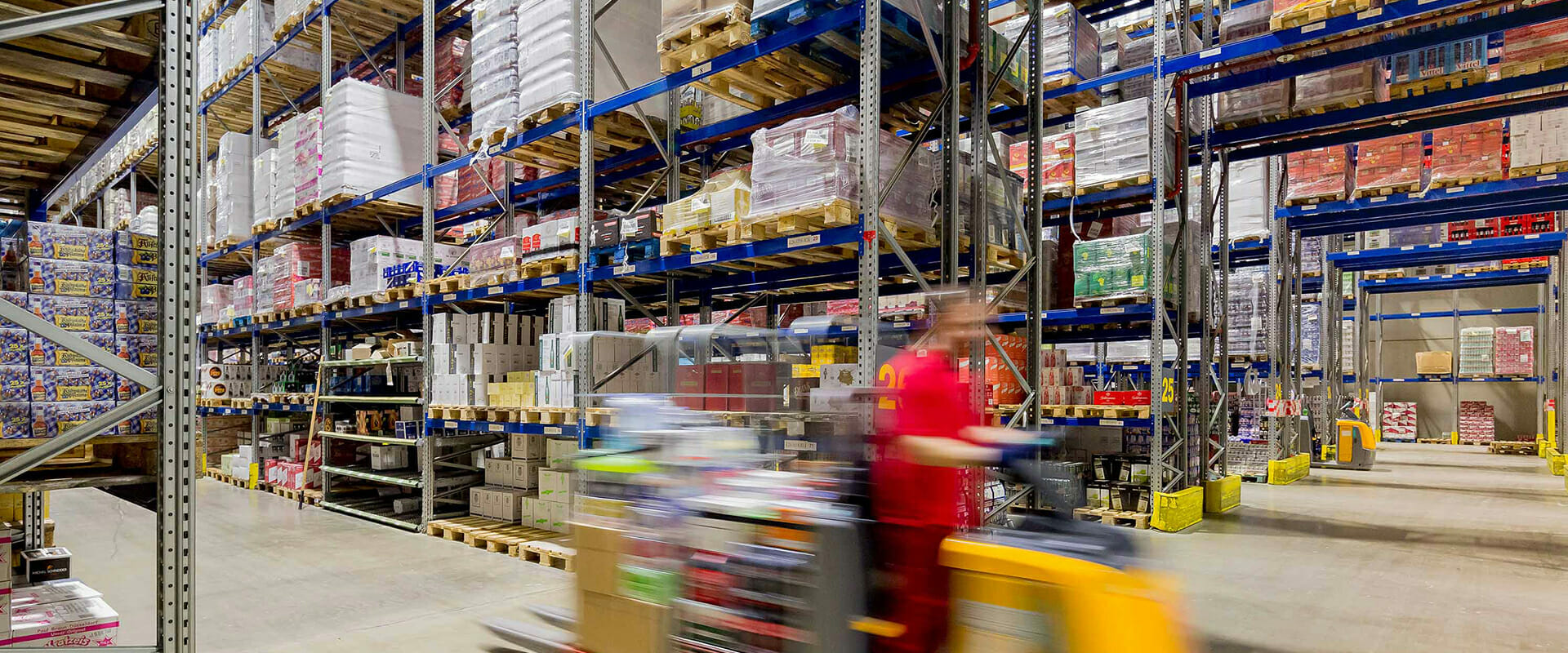 A Lekkerland warehouse with tall shelves storing various goods. A worker in motion operates a pallet jack in the foreground, fulfilling online wholesale orders.