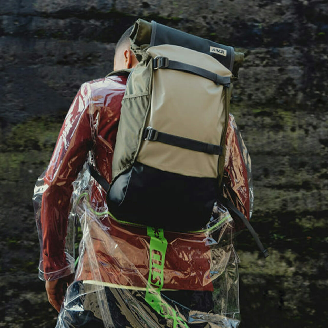 Person wearing a clear plastic raincoat and carrying a large beige and black backpack with a green strap, walking outdoors, as if engaged in step-by-step replatforming of their journey.