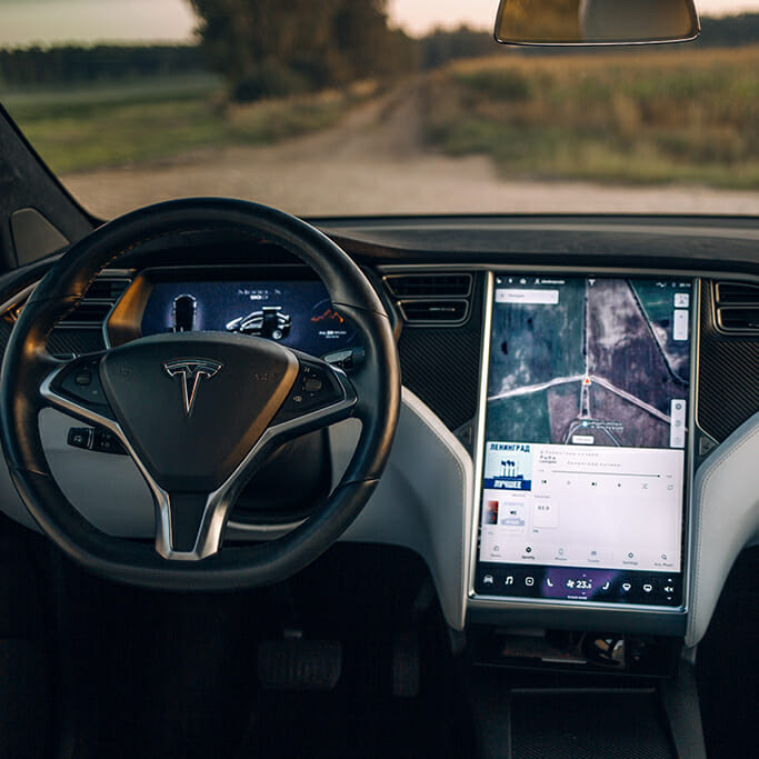 Interior of a car, showcasing a Tesla steering wheel and a large central touchscreen with ERP-integrated navigation and media controls, while an outdoor path is visible through the windshield.