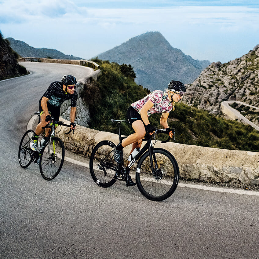 Two cyclists ride uphill on a winding mountain road, with one in the lead. Both wear helmets and cycling gear from ROSE Bikes. Rugged terrain and mountainous landscape are visible in the background.