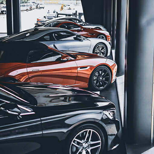 A row of parked luxury sports cars, with a sleek black car in the foreground, followed by red, silver, and white cars inside a modern showroom. The display is so meticulously organized it's as if an ERP system were managing the arrangement.