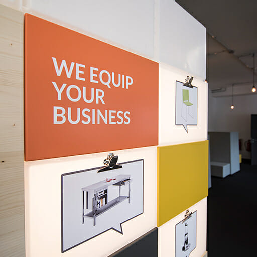 A wall display from Certeo with the text "We Equip Your Business" and images of equipment. The display features a combination of orange, white, and yellow panels, perfect for B2B procurement environments.