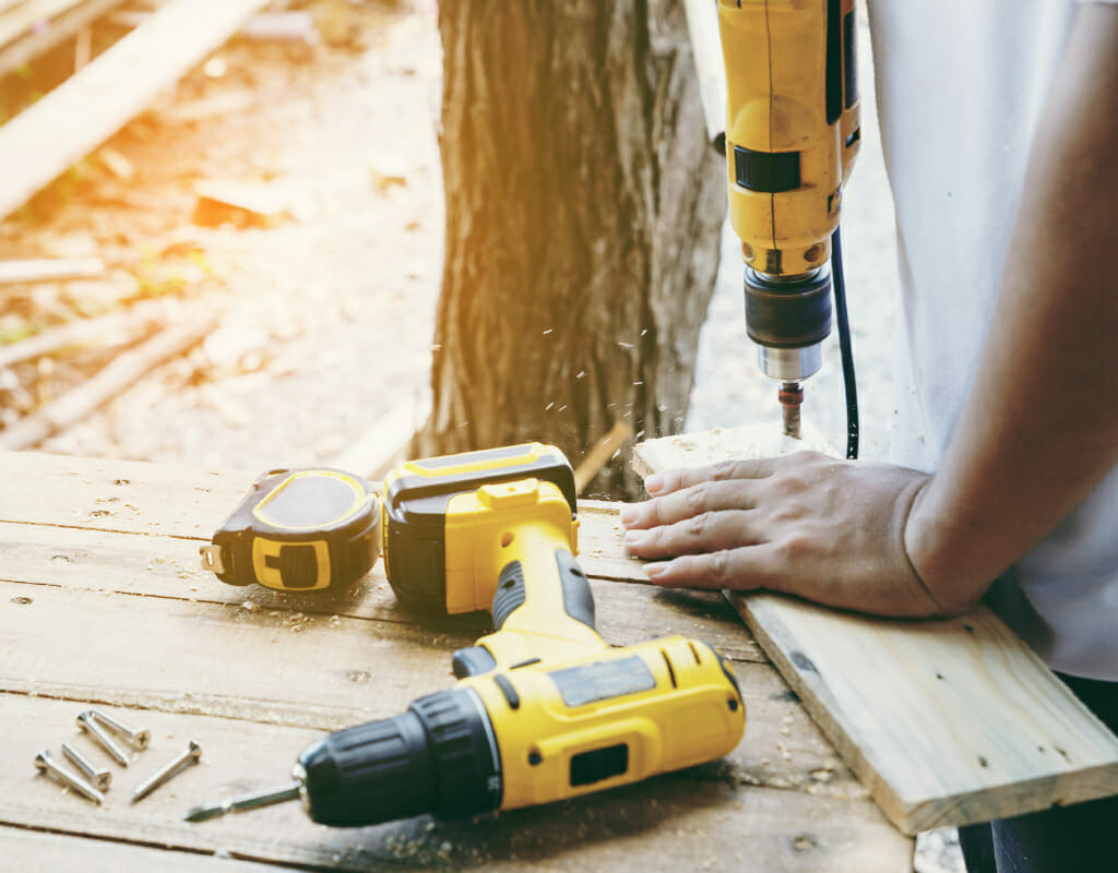 A person uses a power drill on a piece of wood at a workbench, surrounded by various tools including a measuring tape and screws, all stored neatly in a jumbo case.