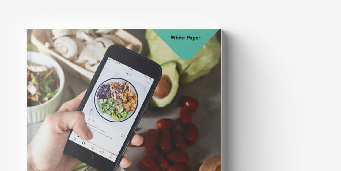A person holding a smartphone proudly displays a food photo on a social media app against a background of fresh vegetables and a printed white paper, highlighting the offerings from an online supermarket.