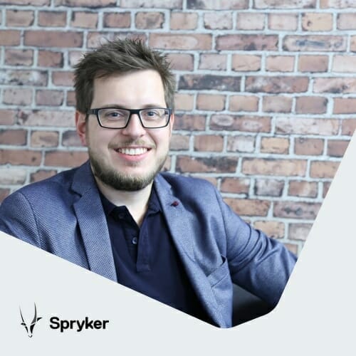 A man with glasses and a beard, wearing a blue blazer over a dark shirt, sits against a brick wall backdrop. The text "Spryker" with a logo appears in the bottom left corner, highlighting the potential of commerce innovations like Click&Collect as retail boosters.