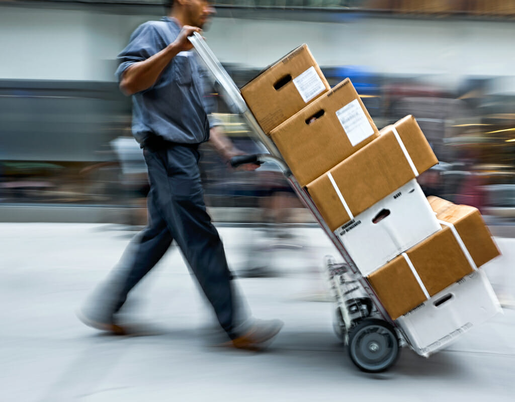 A person is pushing a hand truck loaded with several cardboard boxes along a busy street. The motion blur suggests the person is moving quickly.