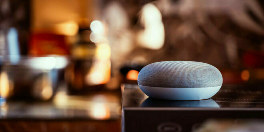 A gray smart speaker sits on a kitchen countertop, seamlessly integrating into the scene with a blurred background of kitchen items and warm lighting. With voice commerce capabilities, it effortlessly assists in ordering groceries and managing daily tasks.