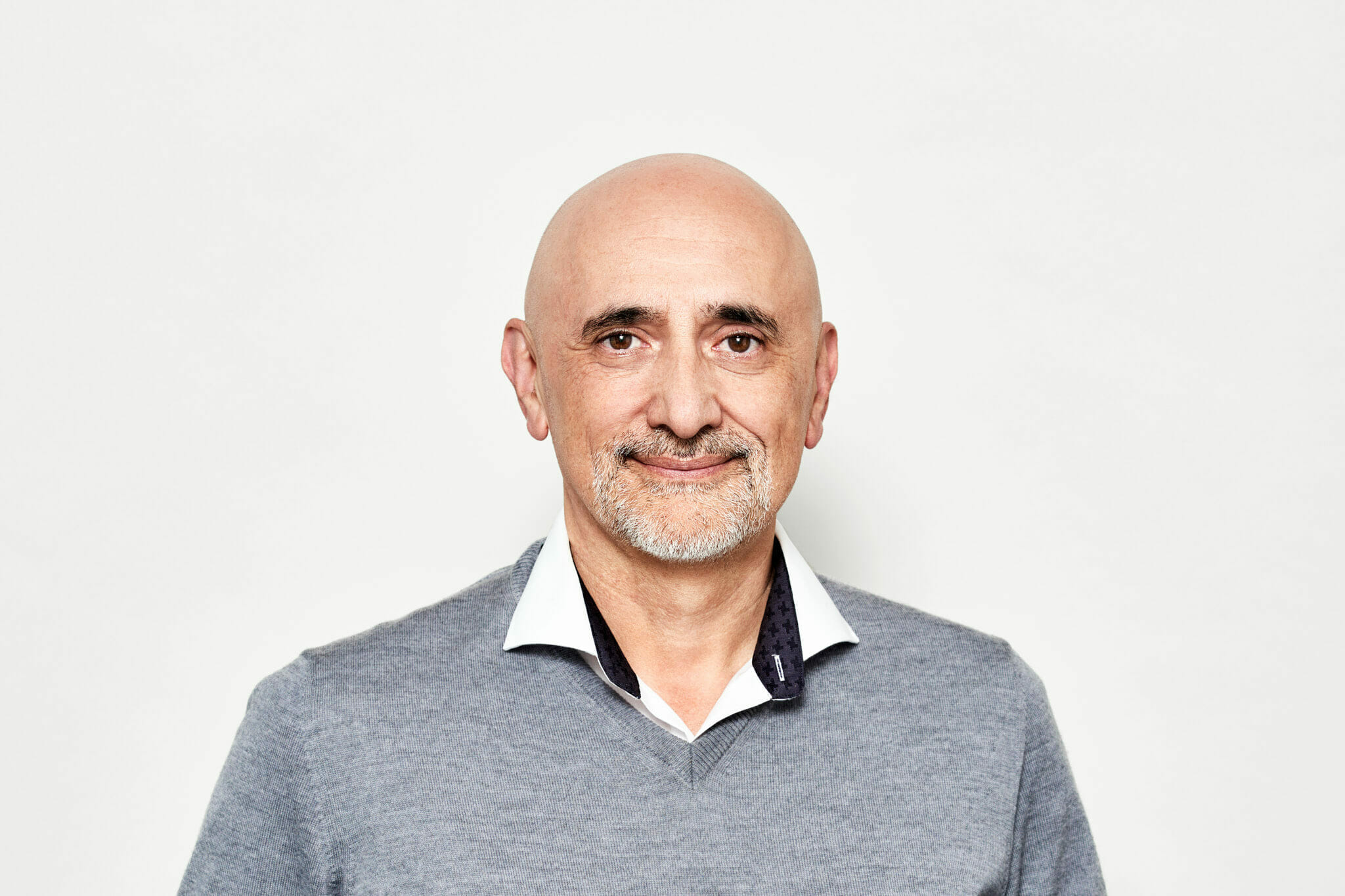 A bald man with a beard and mustache, wearing a gray sweater and a white-collared shirt, stands against a plain white background. He appears to be getting ready for an EXCITE event hosted by Spryker.