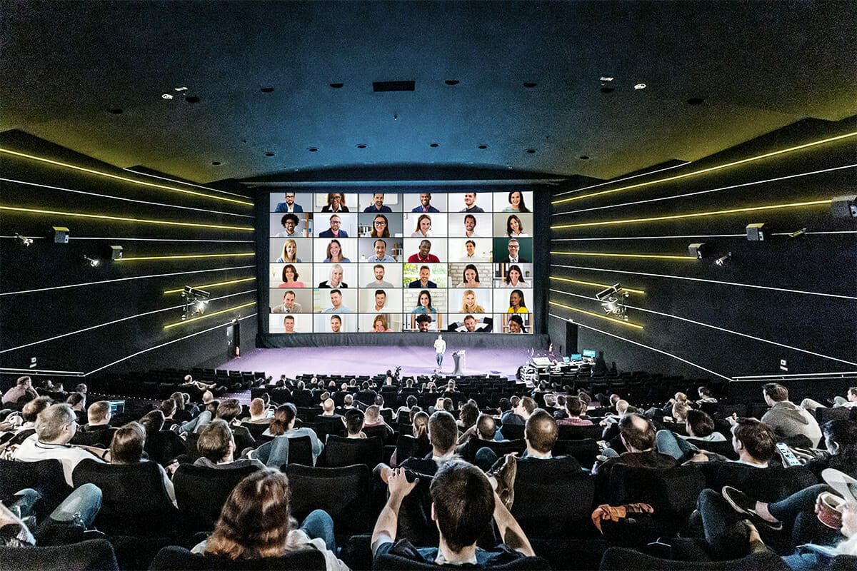 A large conference room with an audience facing a screen displaying multiple people's faces in a video call. A person stands on stage addressing the attendees at Spryker EXCITE.