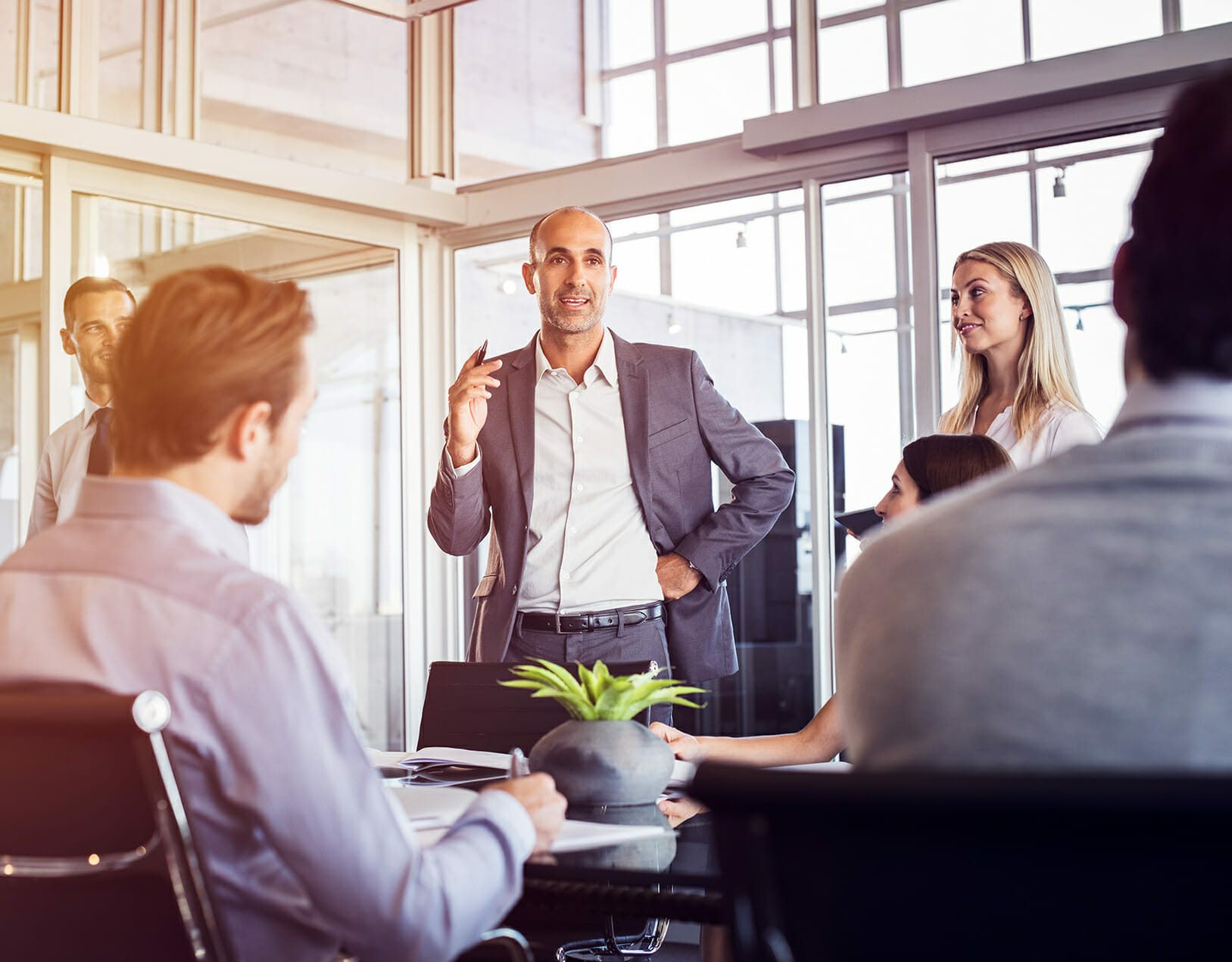 A bald man in a suit speaks to colleagues during a meeting in a glass-walled office. A woman and other people, possibly discussing IDC's latest trends, listen attentively around a table with documents and a small plant.