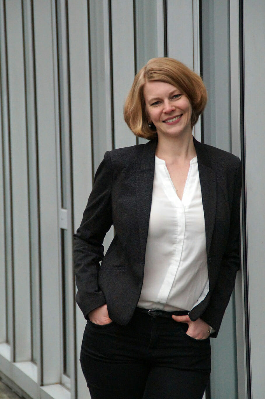 A woman with short blonde hair is smiling, wearing a black blazer over a white blouse, and standing against a modern, vertical-slat wall, ready to go on air.