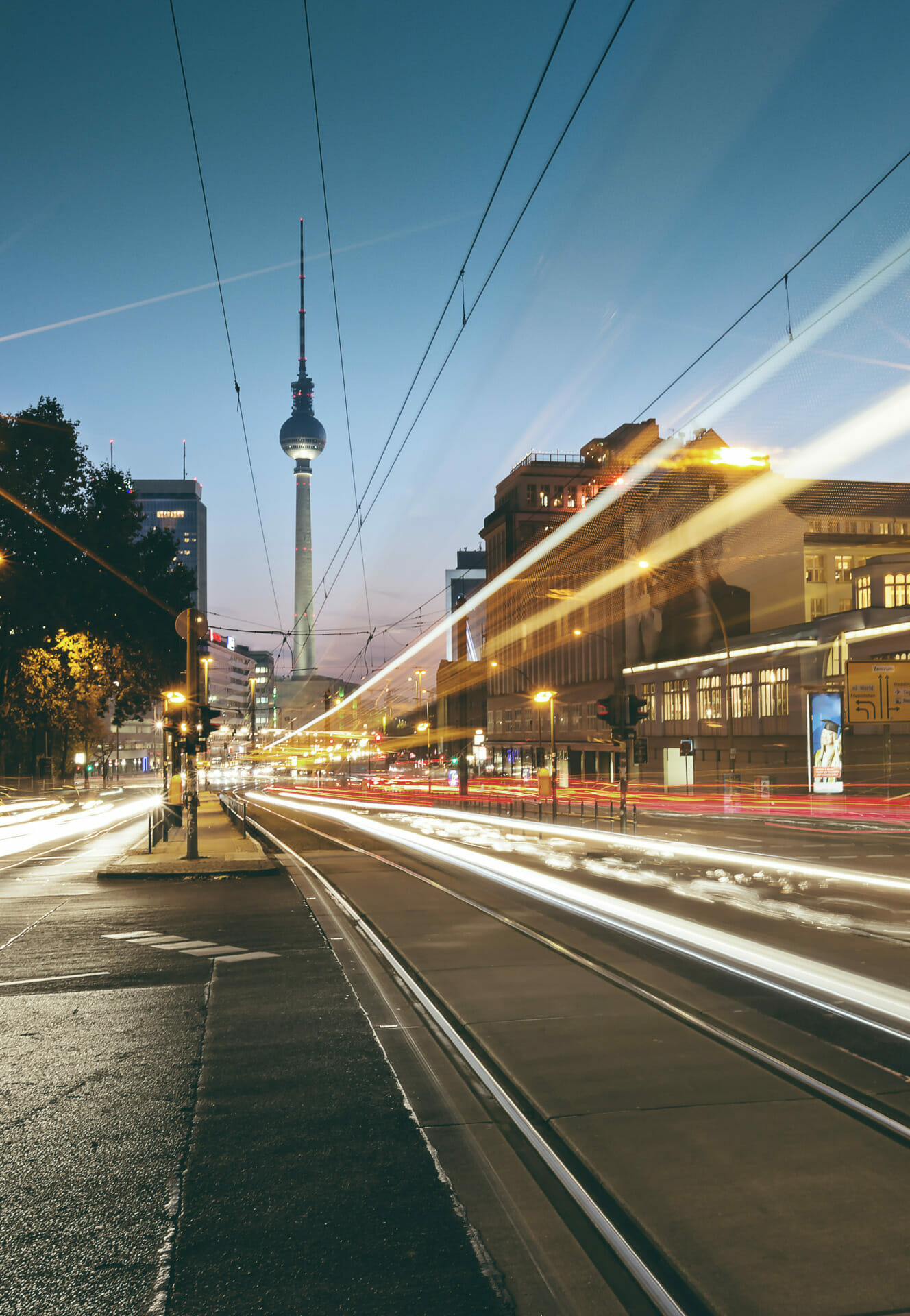 A busy city street at dusk with light trails from cars and trams, buildings illuminated as if celebrating a successful Finanzierungsrunde, and a tall tower visible in the background.