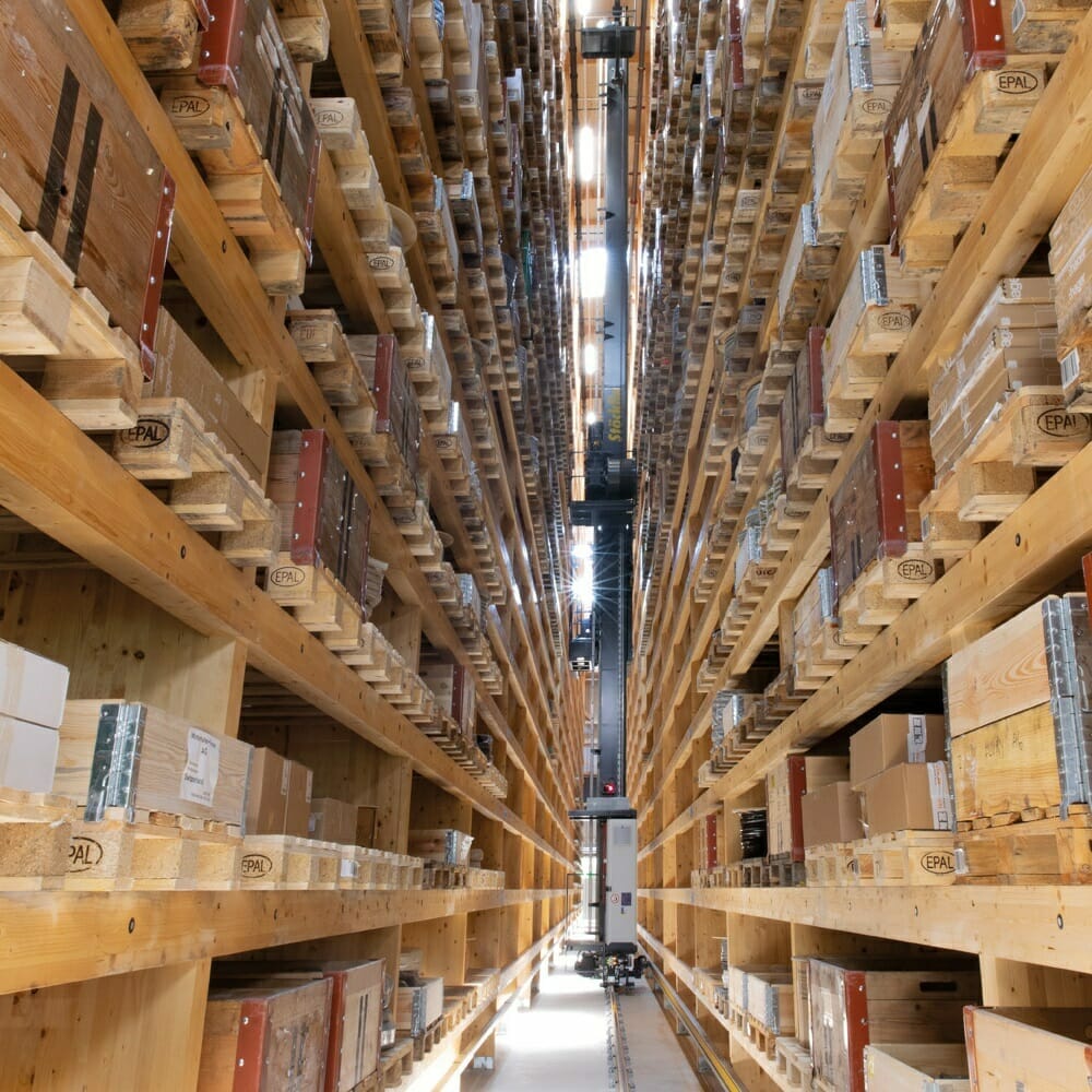 A high-ceiling warehouse aisle with numerous wooden shelves filled with various boxes, a state-of-the-art Winterhalter+Fenner robotic system on tracks in the center aisle.