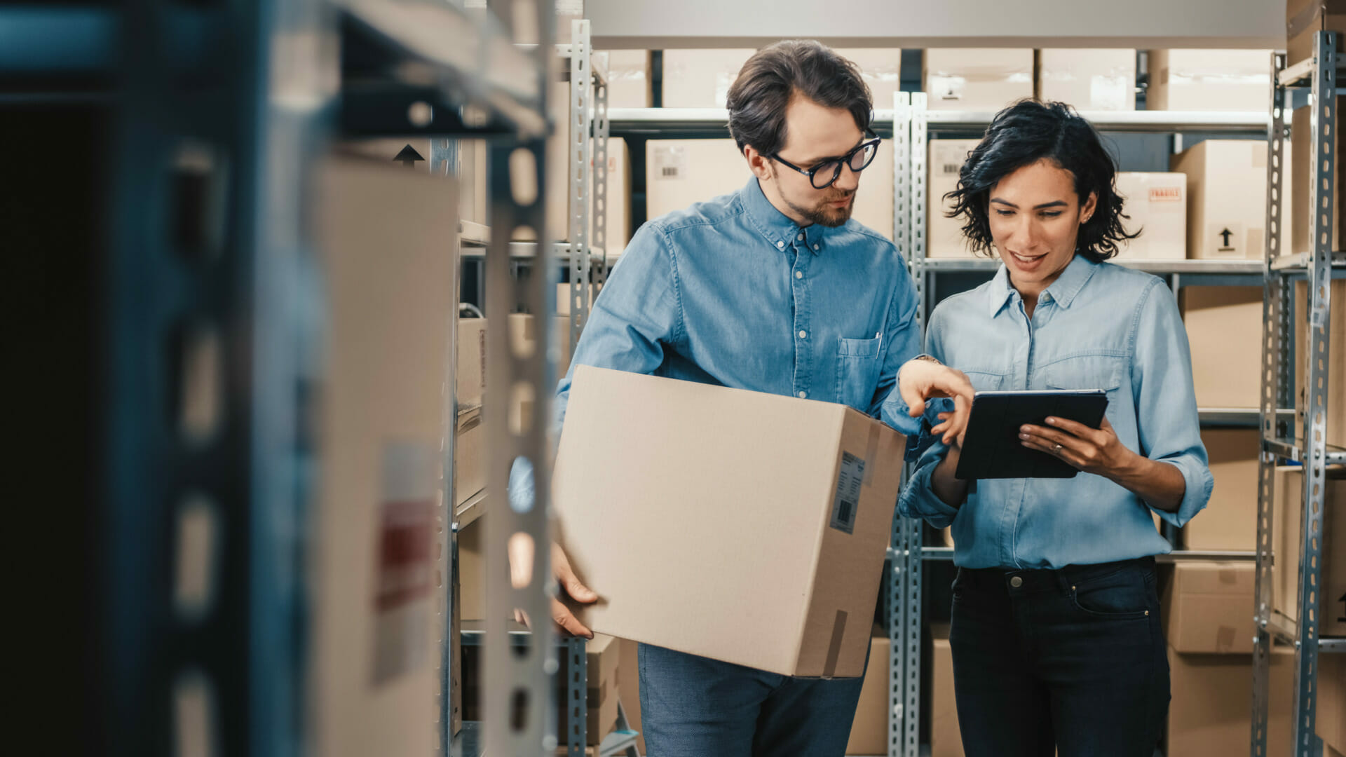 Two people in a storage room, one holding a cardboard box and the other using a tablet to check customer details. Shelves filled with boxes surround them, creating an organized metro of inventory.