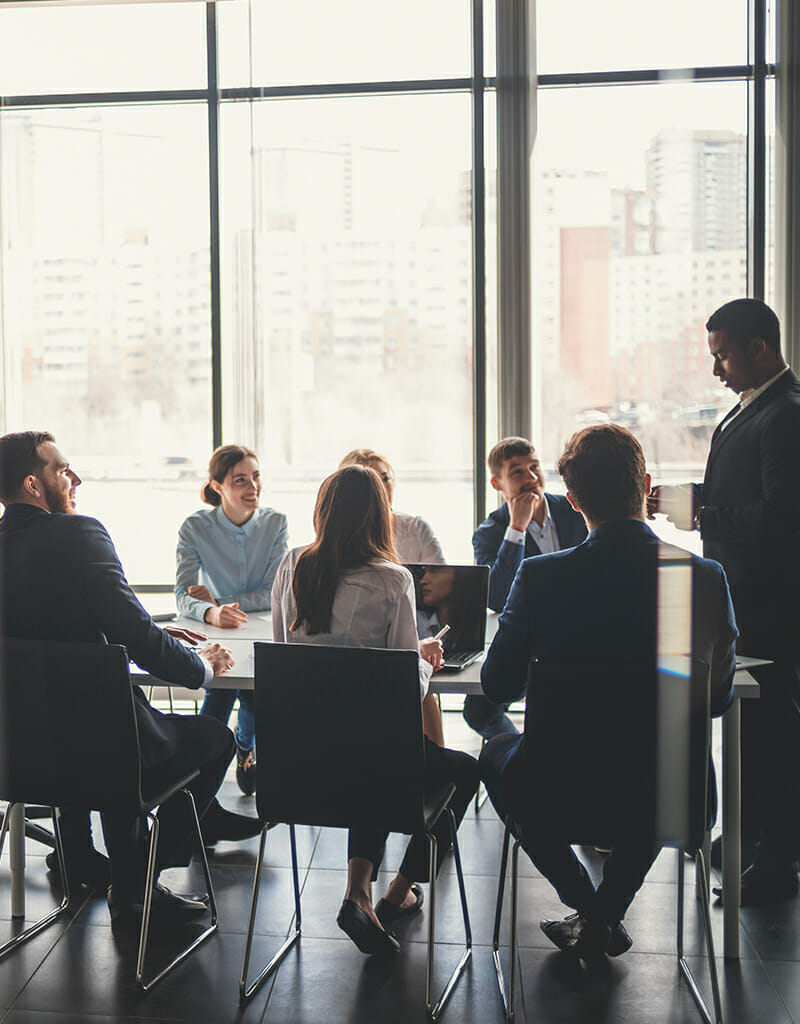 A group of eight people in business attire are seated around a conference table, engaged in discussion. One person is standing and presenting to the group, a project led by Marco Knecht for Spryker OnAir. Large windows in the background show a cityscape.