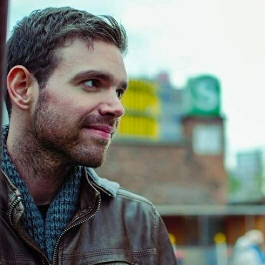 A man with short brown hair and a beard, wearing a brown leather jacket and gray scarf, stands outdoors with a blurred building in the background, discussing the potential of voice commerce.