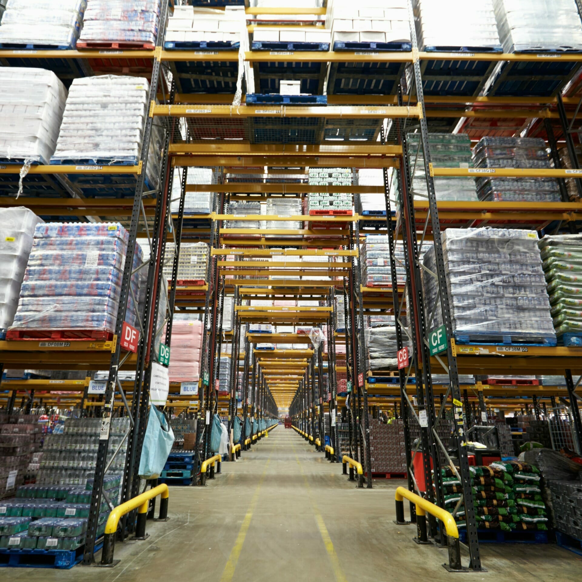 A large warehouse aisle with tall racks filled with various packaged goods, including beverages and other consumables from Stauff. Yellow guardrails line the racks, and the floor is clear for transportation.