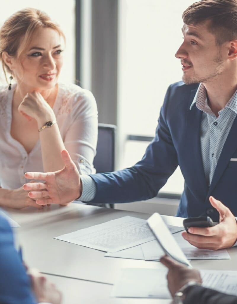 Two people in professional attire engage in a discussion at a meeting table, with documents and a tablet in front of them, possibly planning for Disaster Nights at Hannover Messe 2024. The man gestures while speaking, and the woman listens attentively.