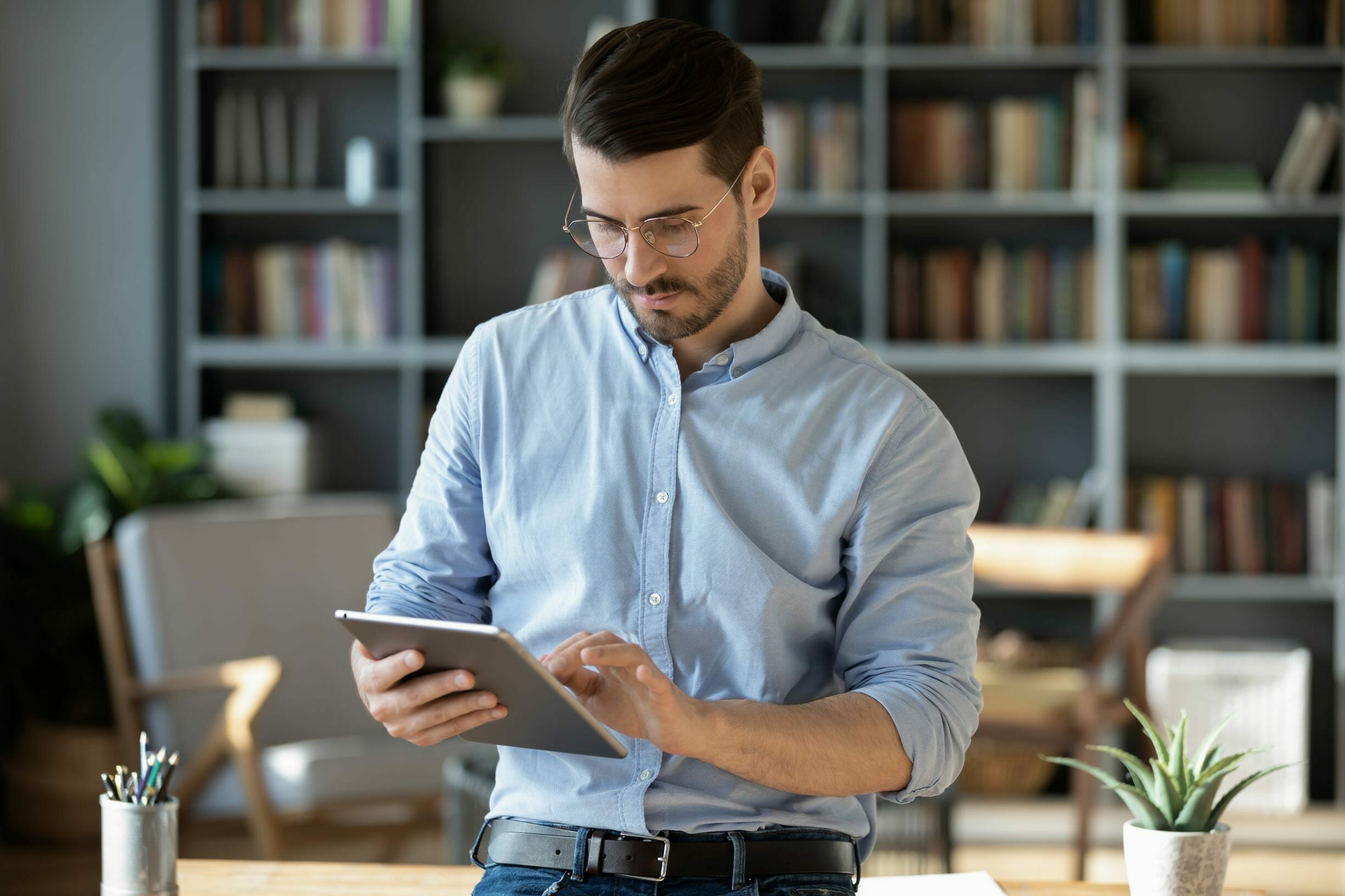 A man with glasses, dressed in a light blue shirt, is using a tablet in a room filled with bookshelves and a desk, likely exploring the latest trends in composable commerce.