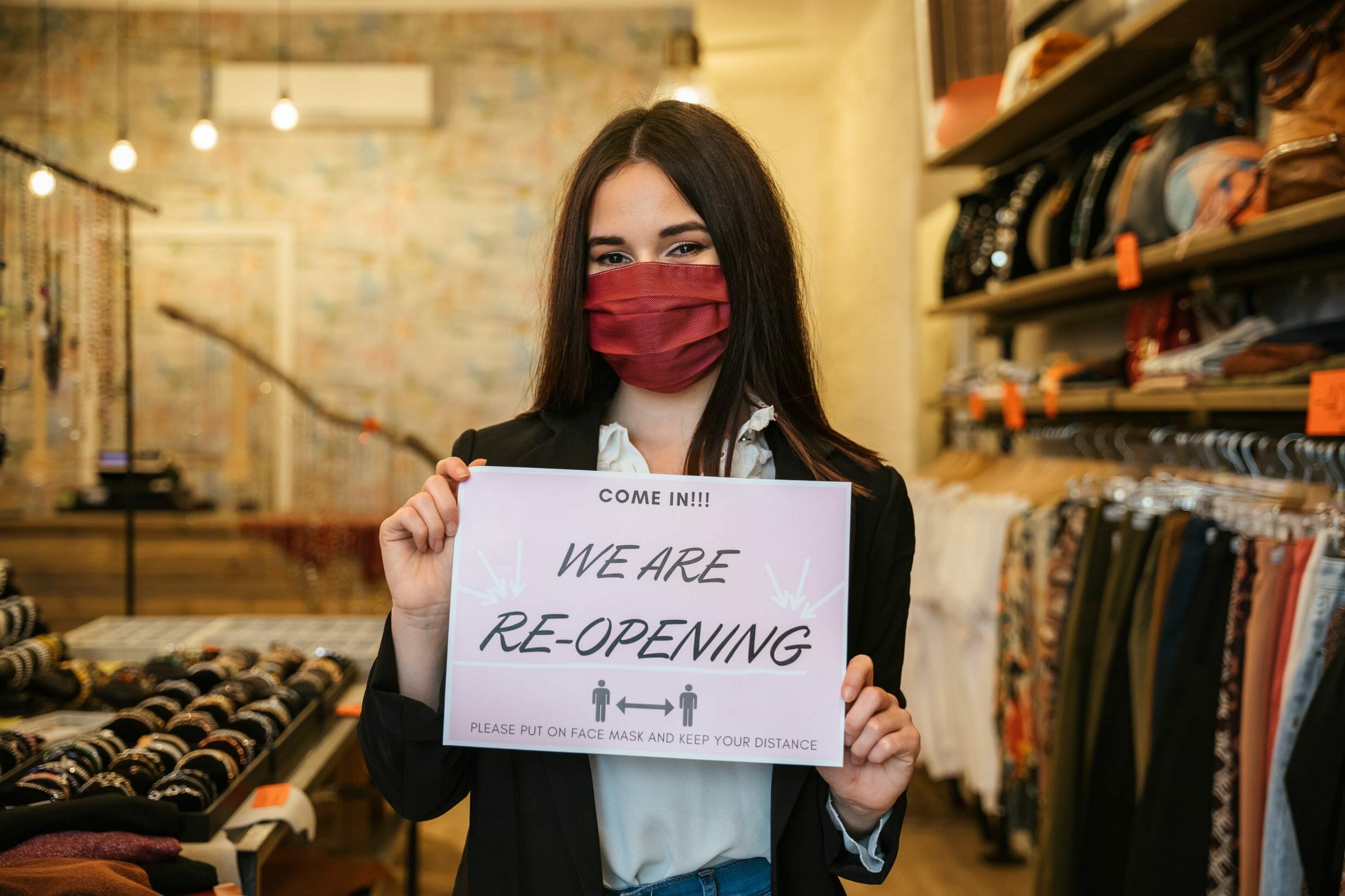 A woman wearing a red face mask and a black jacket holds a sign in a clothing store that says "We are re-opening. Please use contactless service, put on your face mask, and keep your distance.