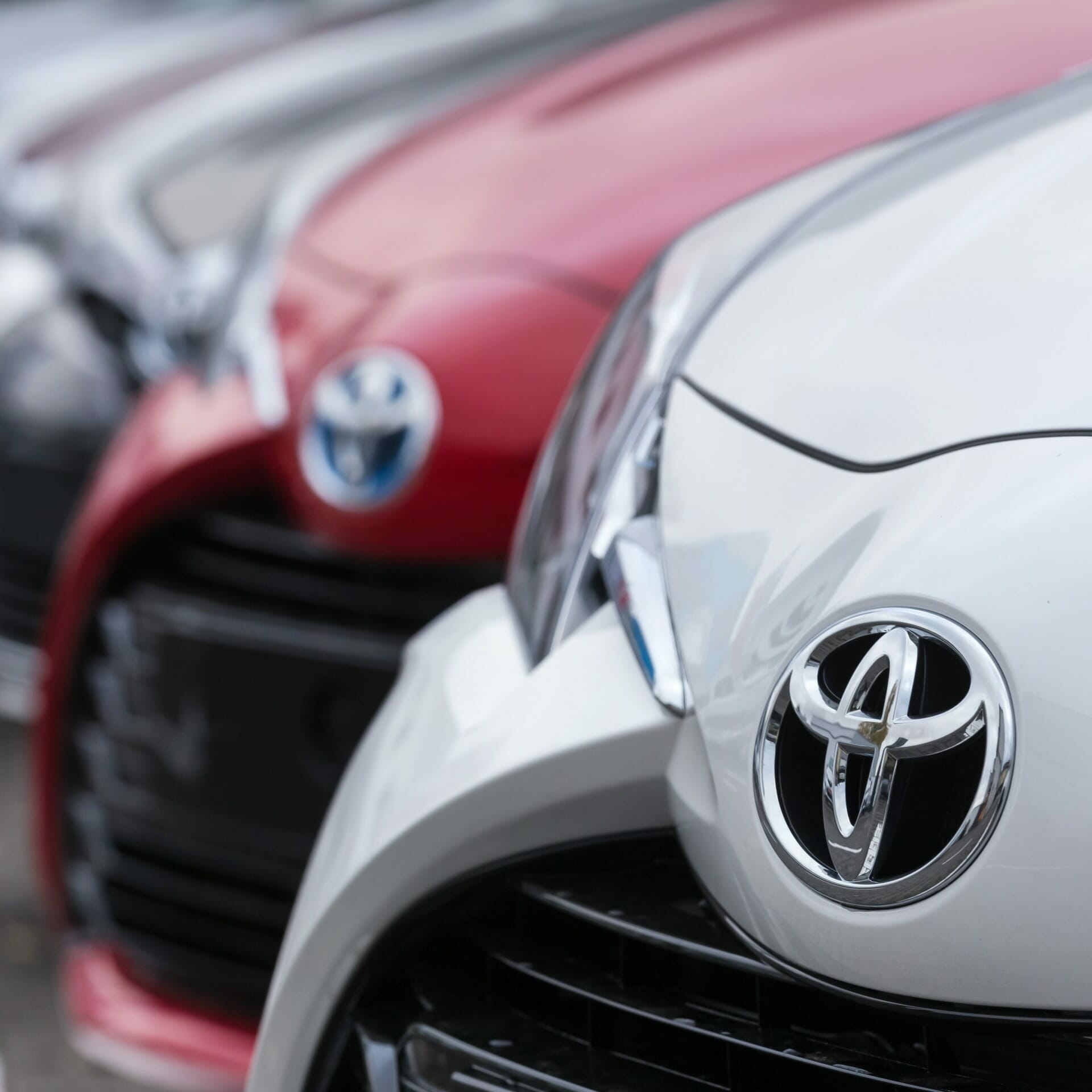 Row of parked Toyota cars with front view of grilles and logos, featuring a white and a red car in the foreground. This display is part of the efficient Toyota shop system.