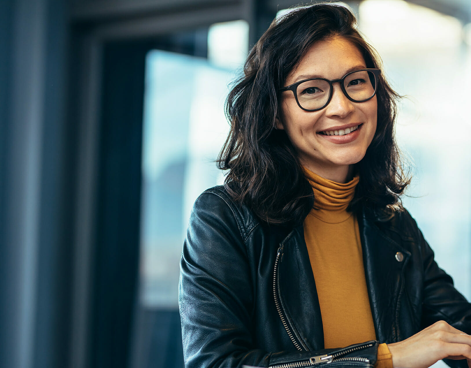 A person with glasses and a yellow turtleneck smiles while wearing a black leather jacket, embodying the confident style of a TrustRadius professional.