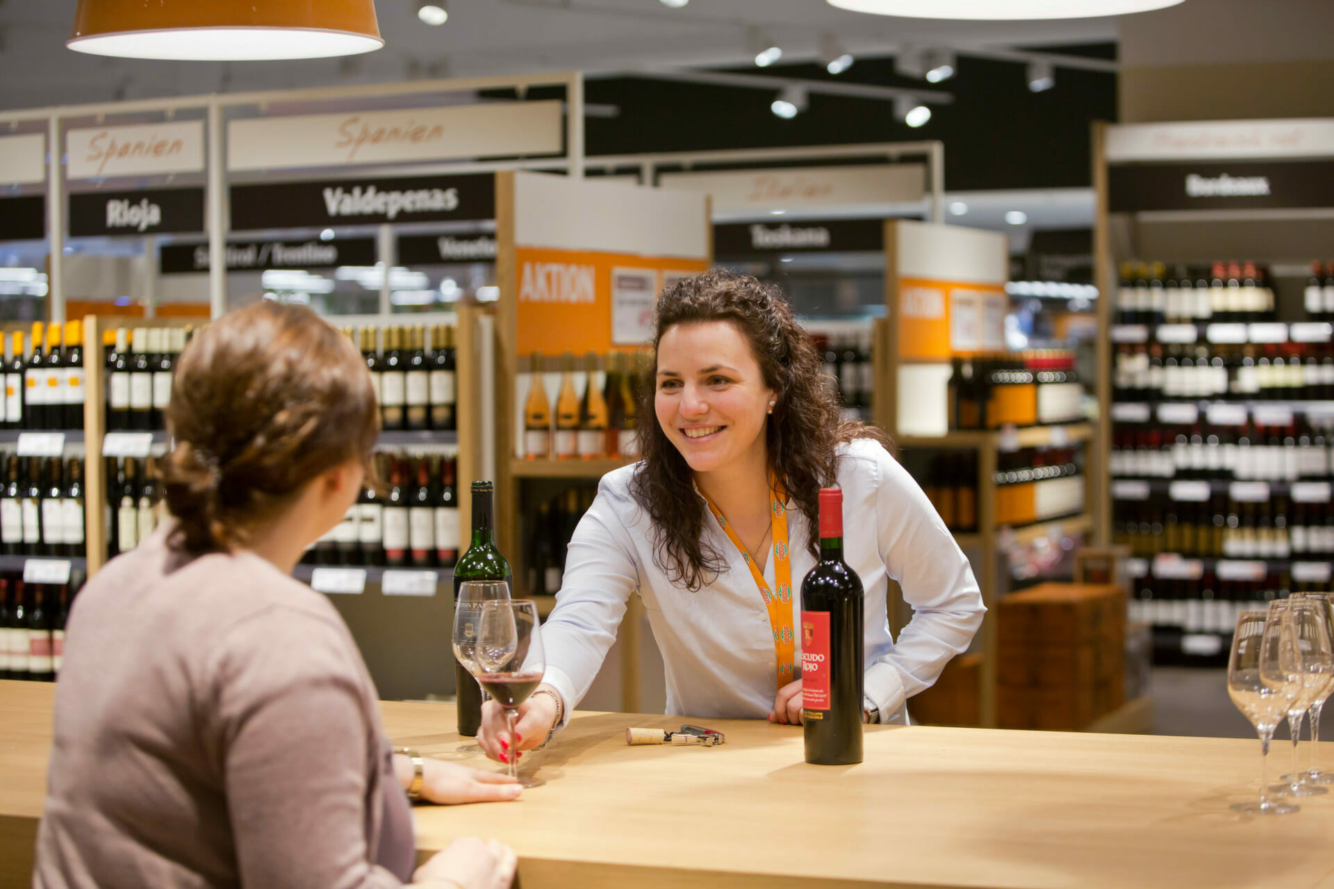 A woman in a wine store pours red wine into a glass for another woman. The store, named Globus, has labeled sections and shelves filled with various wine bottles.