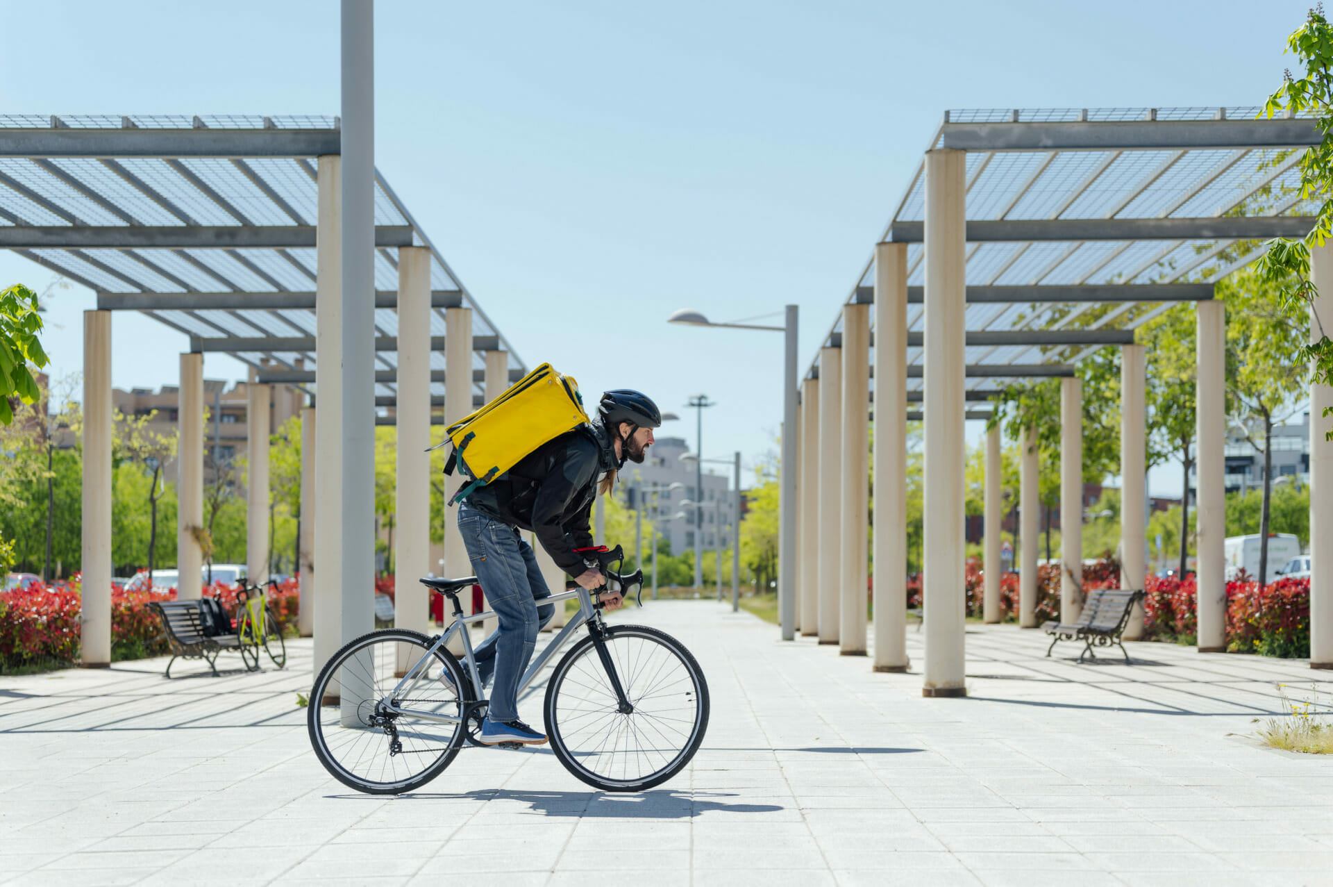 A person wearing a helmet and backpack, evoking the spry energy of Spryker OnAir, is riding a bicycle along a paved path with wooden pergolas and greenery on either side.