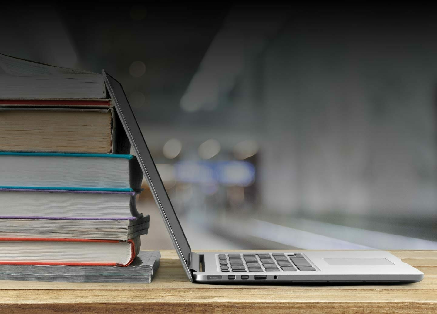 A laptop partially open next to a stack of books on a wooden table with a blurred background, symbolizing the blend of eCommerce tech and academic pursuits typical of Spryker Academy.