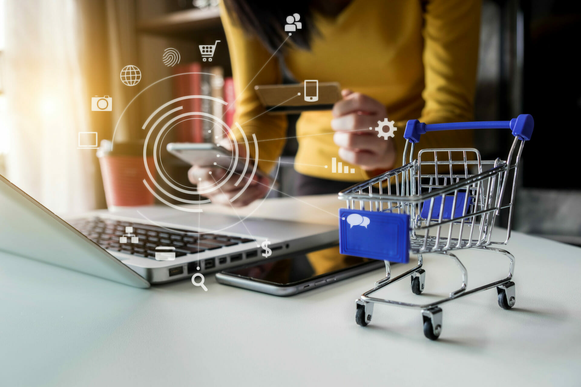 A person using a smartphone and holding a credit card in front of a laptop, with a small shopping cart model nearby and digital icons representing online shopping and technology overlayed, showcases the seamless B2C user experience.