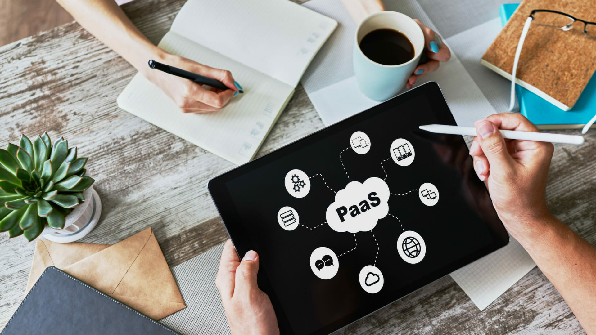 Two people work at a desk with notebooks, a tablet displaying a "PaaS" diagram, a coffee cup, and a potted plant. One person writes while the other points at the tablet with a stylus, providing support as they collaborate.