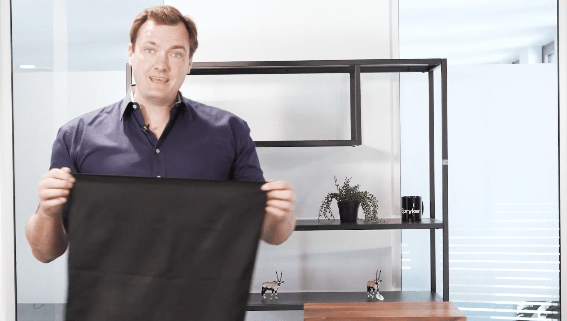 A man in a blue shirt holds a black rectangular cloth, standing indoors with a black shelving unit and small plants behind him, reminiscent of an e-commerce setup with a touch of magic.