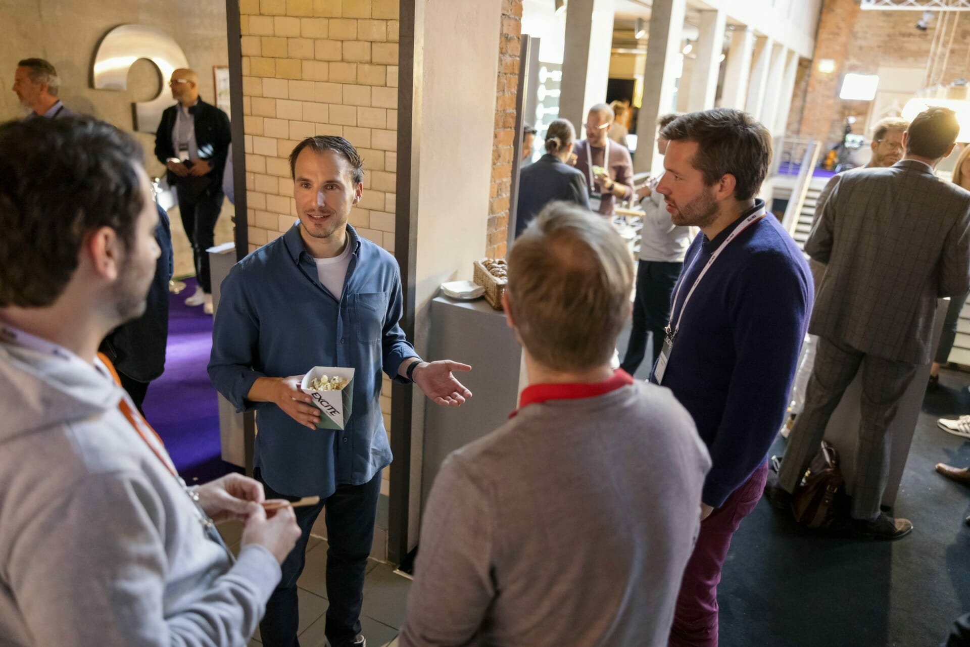 A group of four men are engaged in conversation at a casual indoor event, reminiscent of a Las Vegas conference atmosphere. One man is holding a popcorn container. Other attendees and an event space are visible in the background, creating an ambiance perfect for Shoptalk discussions.