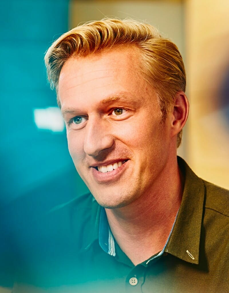 A smiling man with short blond hair is looking slightly to the right, with a blurred blue and yellow background. This could be Guido Jansen in a candid moment.