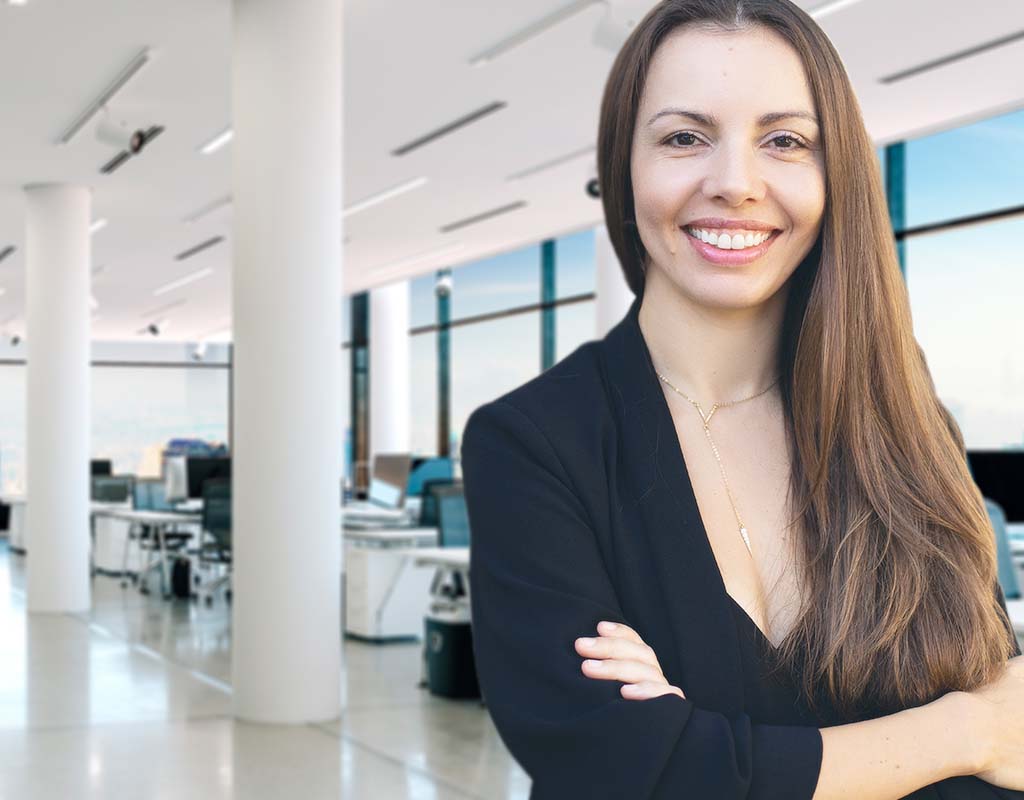 Elena Leonova, a woman with long brown hair and a black outfit, stands smiling in a modern, well-lit office with large windows and white pillars.