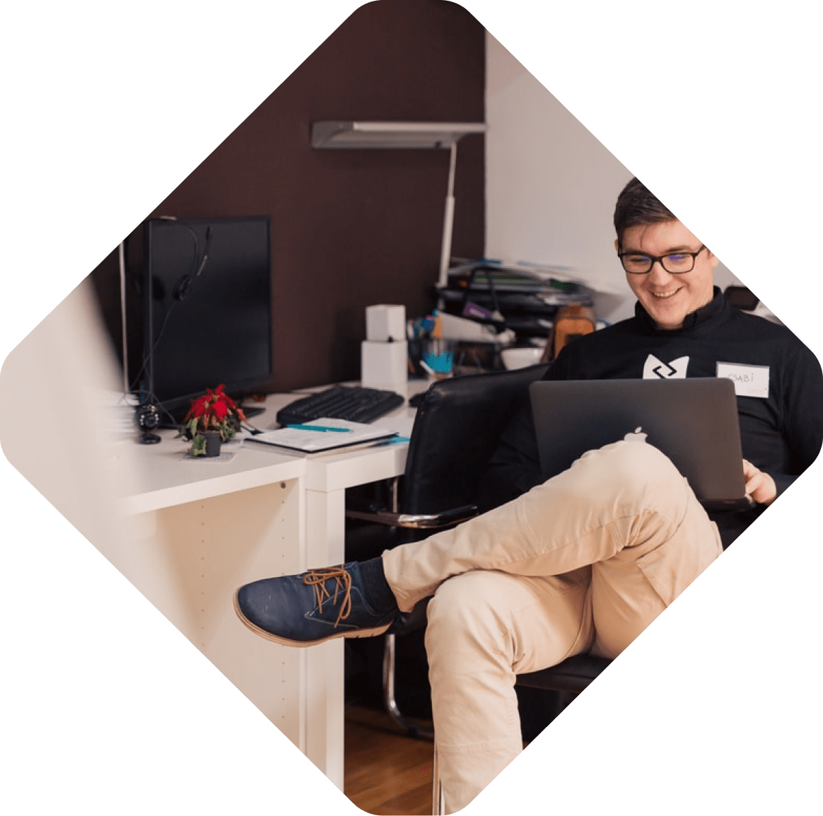 A person sits in an office chair, smiling while using a laptop. To their left, a desk holds a computer monitor, keyboard, and small potted plant. They are researching composable commerce strategies with enthusiasm evident on their face.