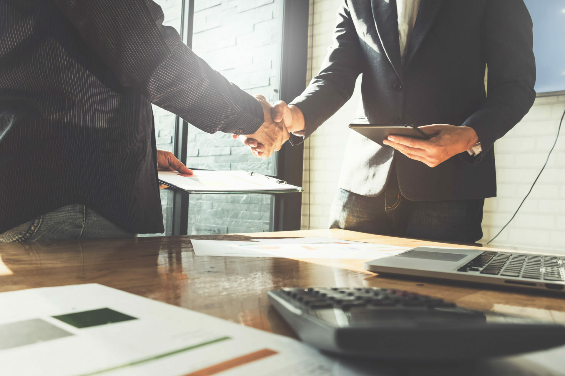 Two business professionals shaking hands over a desk with documents, a calculator, laptop, and tablet in view, as if finalizing press releases. Sunlight streams through the window in the background.