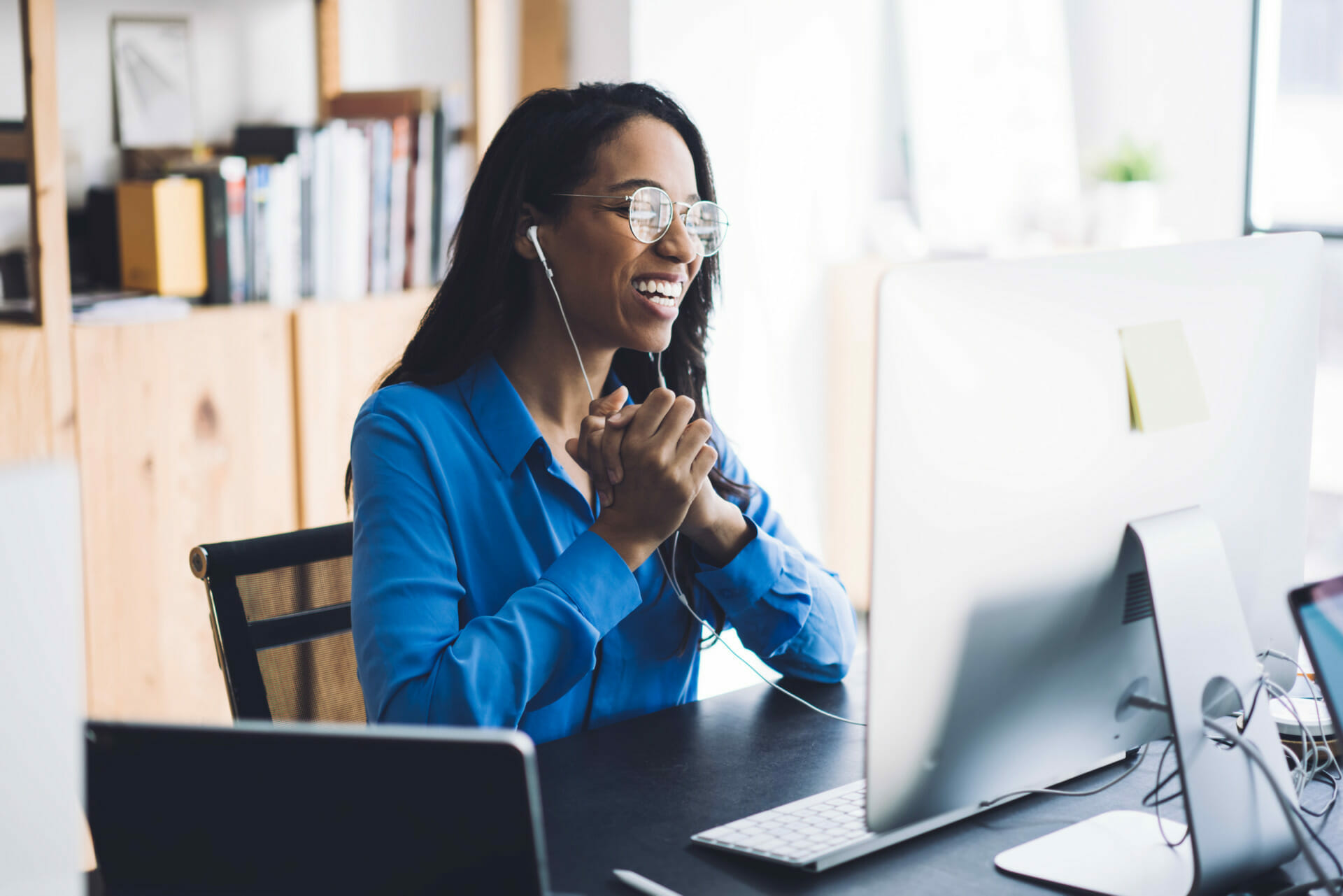 A woman wearing glasses and earphones smiles while sitting at her desk, engrossed in a Forrester report on her computer screen.