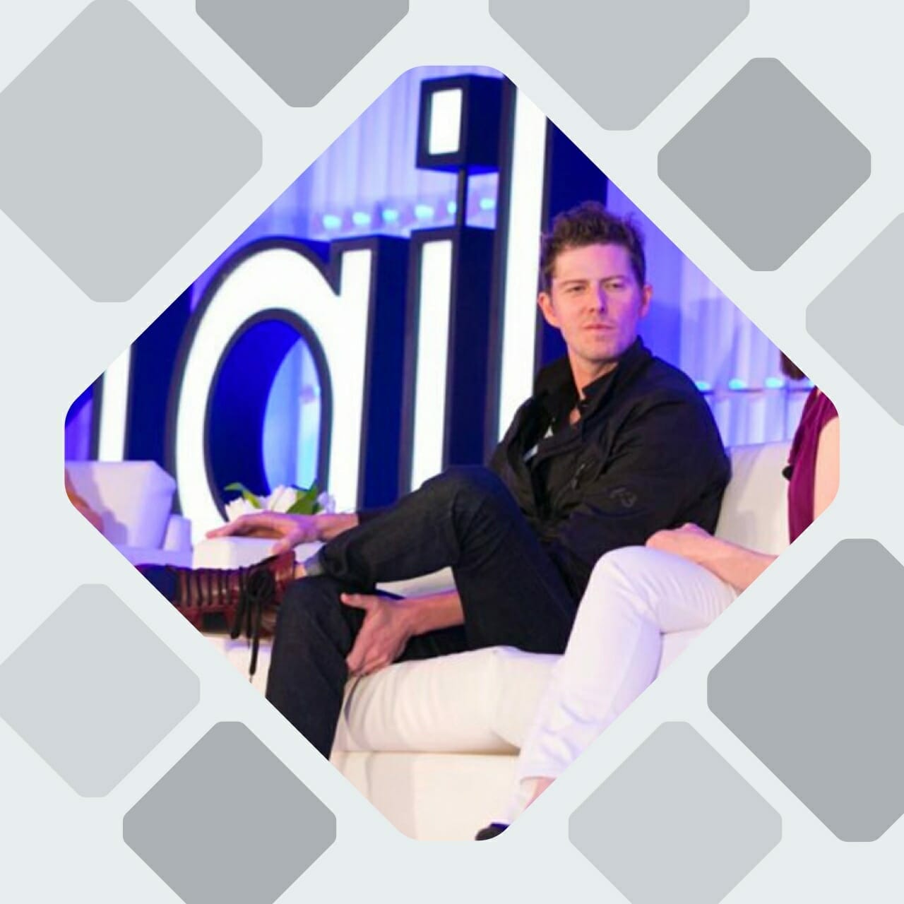 A man in a black shirt sits on a white couch on a stage, participating in a discussion about e-commerce. Large text behind him spells part of the word "eTail," highlighting the event's focus on online retail advancements.