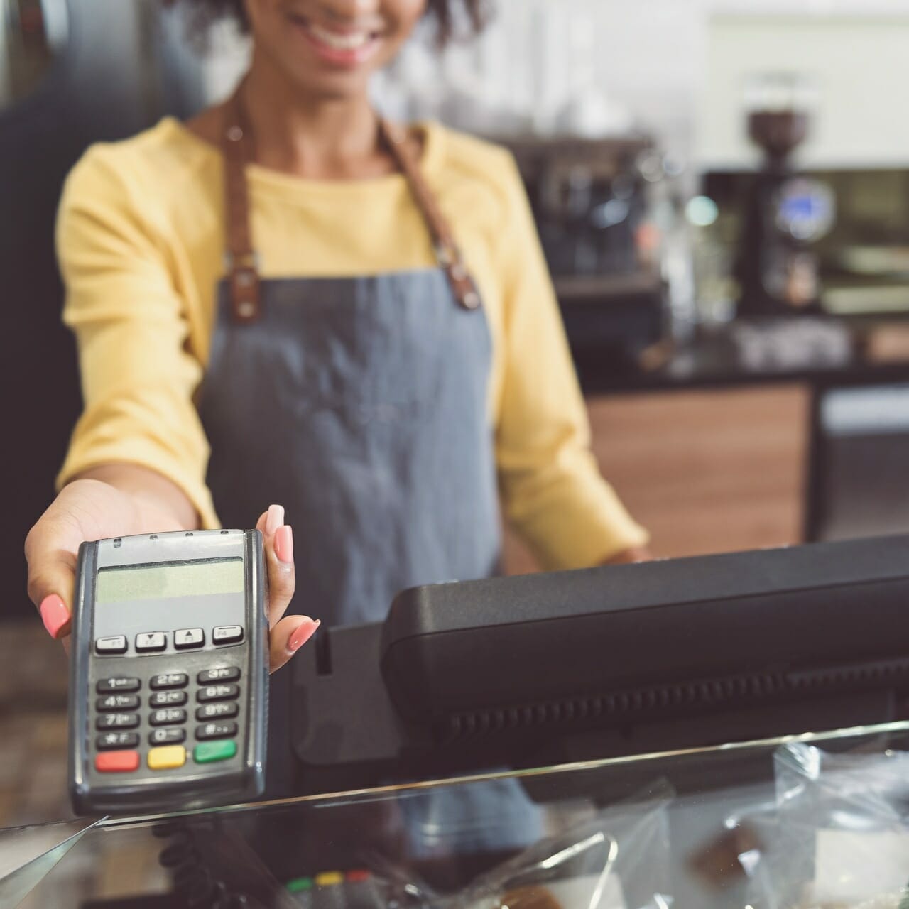 A person wearing an apron is holding a card payment terminal, extending it towards the camera, ready to accept payment. A cash register is on the counter beside them. Join Spryker for ShoppingTomorrow and simplify your retail transactions effortlessly.