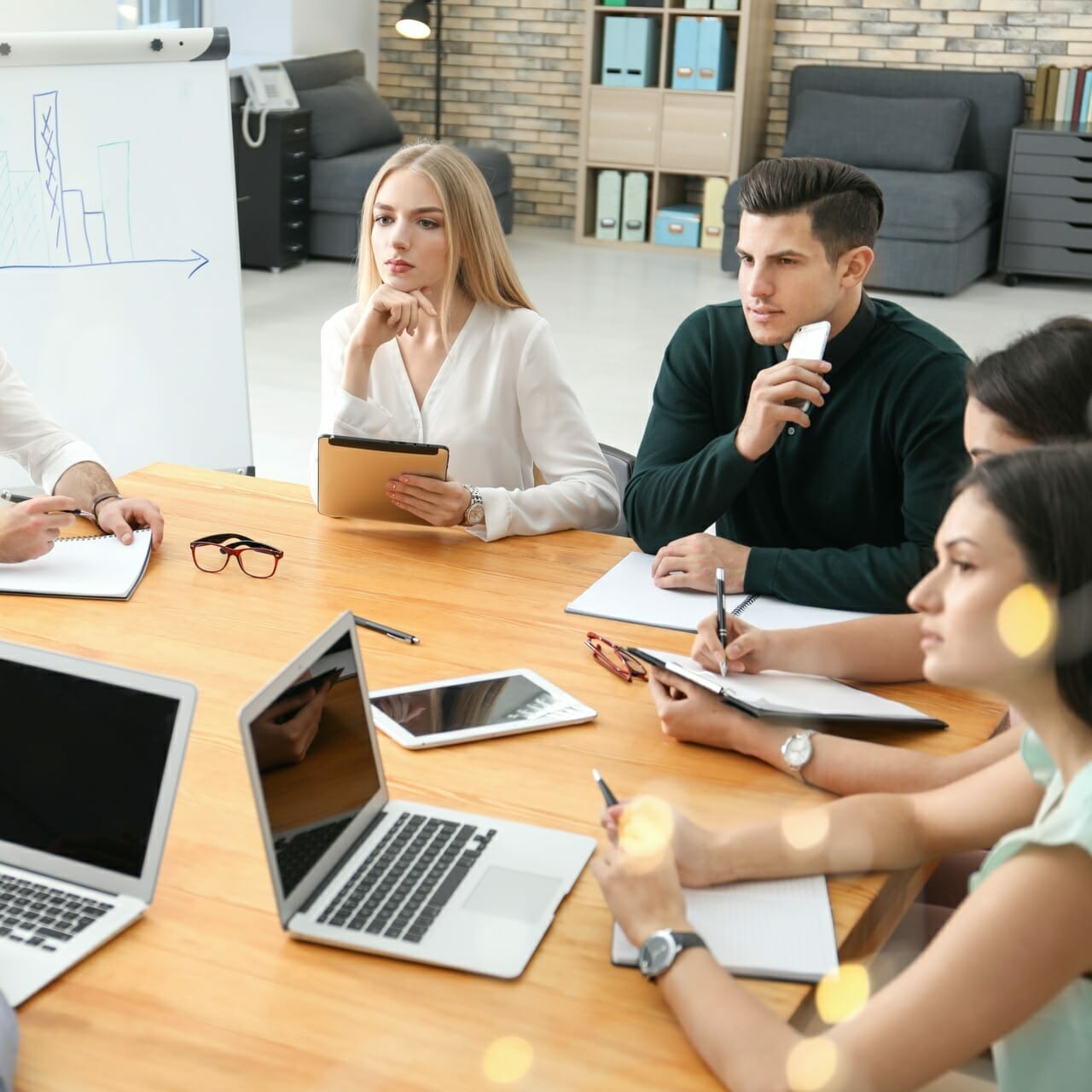 A group of six people are sitting around a table in an office, engaged in a meeting. Laptops, notebooks, and a chart on an easel for the ShoppingTomorrow project are visible. Some are taking notes, while one person holds a tablet displaying Spryker's latest updates.