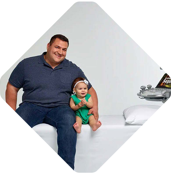 A man sits on a bench next to a smiling baby girl in a green dress and headband, while Siemens Healthineers medical equipment is visible near the bench.