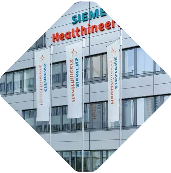 A modern building with the Siemens Healthineers logo displayed at the top. Three flagpoles in front of the building hold white flags, each with the Siemens Healthineers logo. Several windows reflect a cloudy sky. The image is rotated to a diamond orientation.