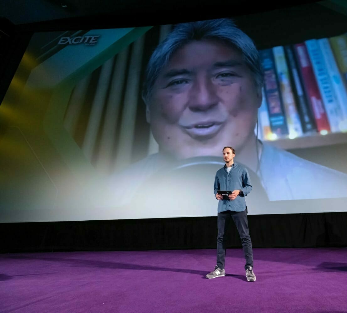 A man stands on a stage with a microphone, addressing an audience. A large screen behind him provides an overview, showing a close-up of another man speaking with books visible in the background.