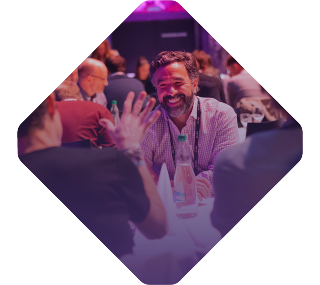 A group of people, including a bearded man smiling, engage in conversation at a table during the Spryker EXCITE 2022 event.