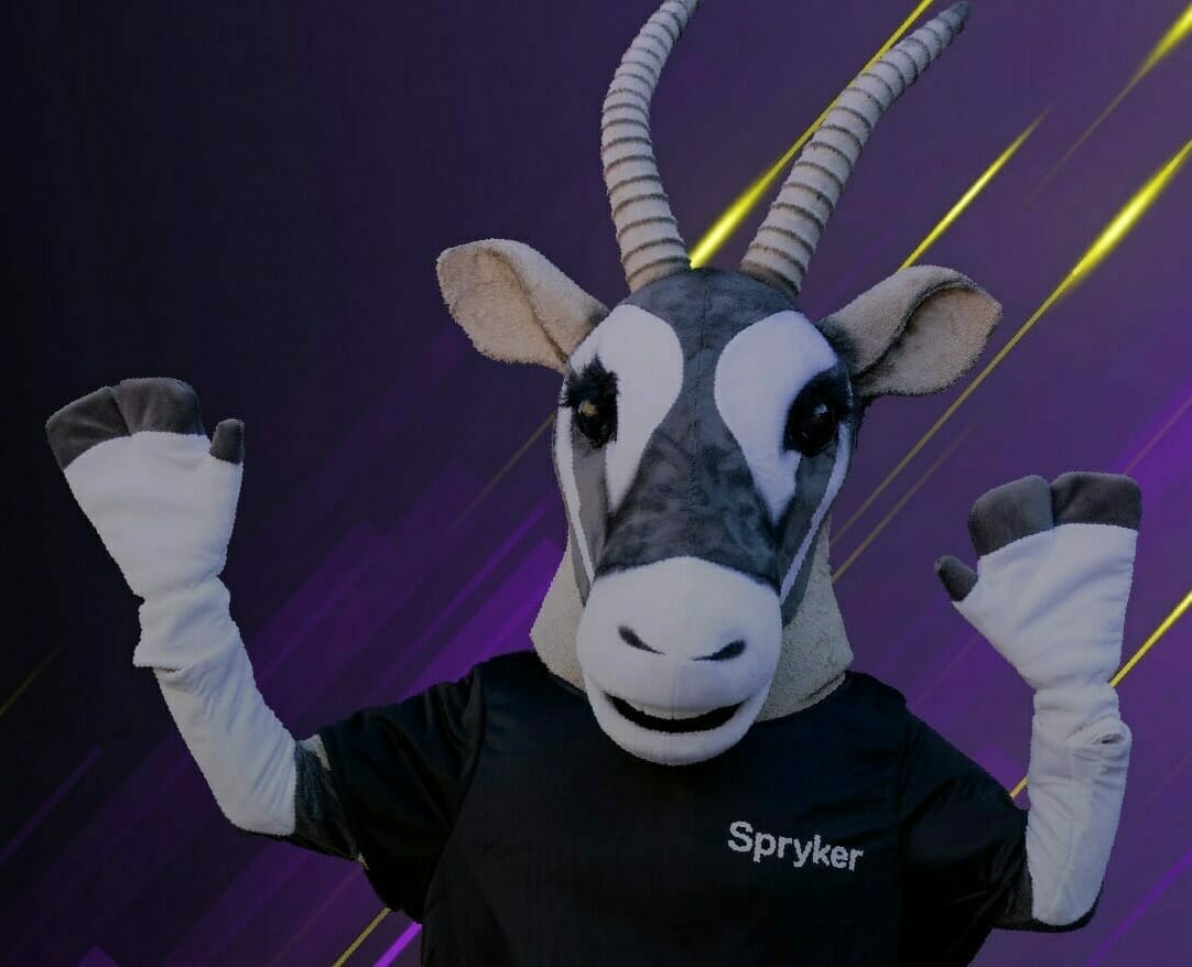 A costumed individual wearing an antelope head with large horns and a black shirt with "Spryker" printed on it, symbolizing e-commerce prowess, stands against a dark purple and yellow background, sporting white gloves.