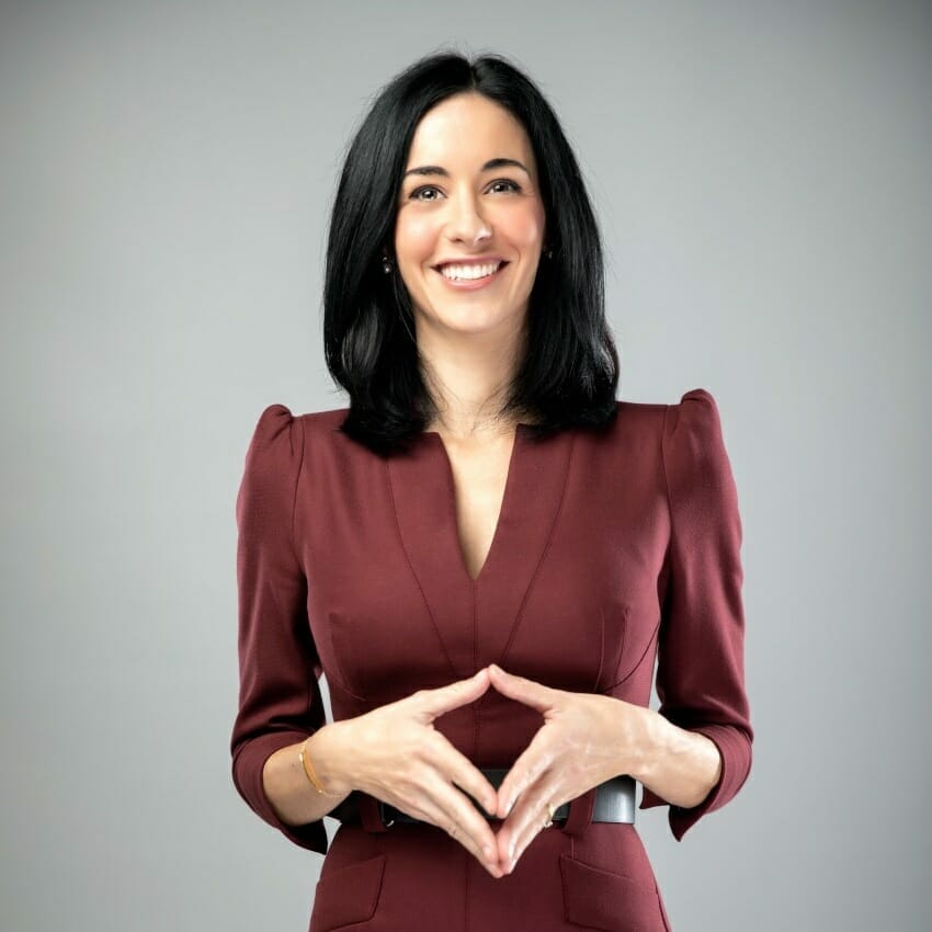 A woman with shoulder-length black hair, wearing a maroon dress with puffed shoulders, stands smiling at the HERd Conference and holding her hands together against a plain gray background.