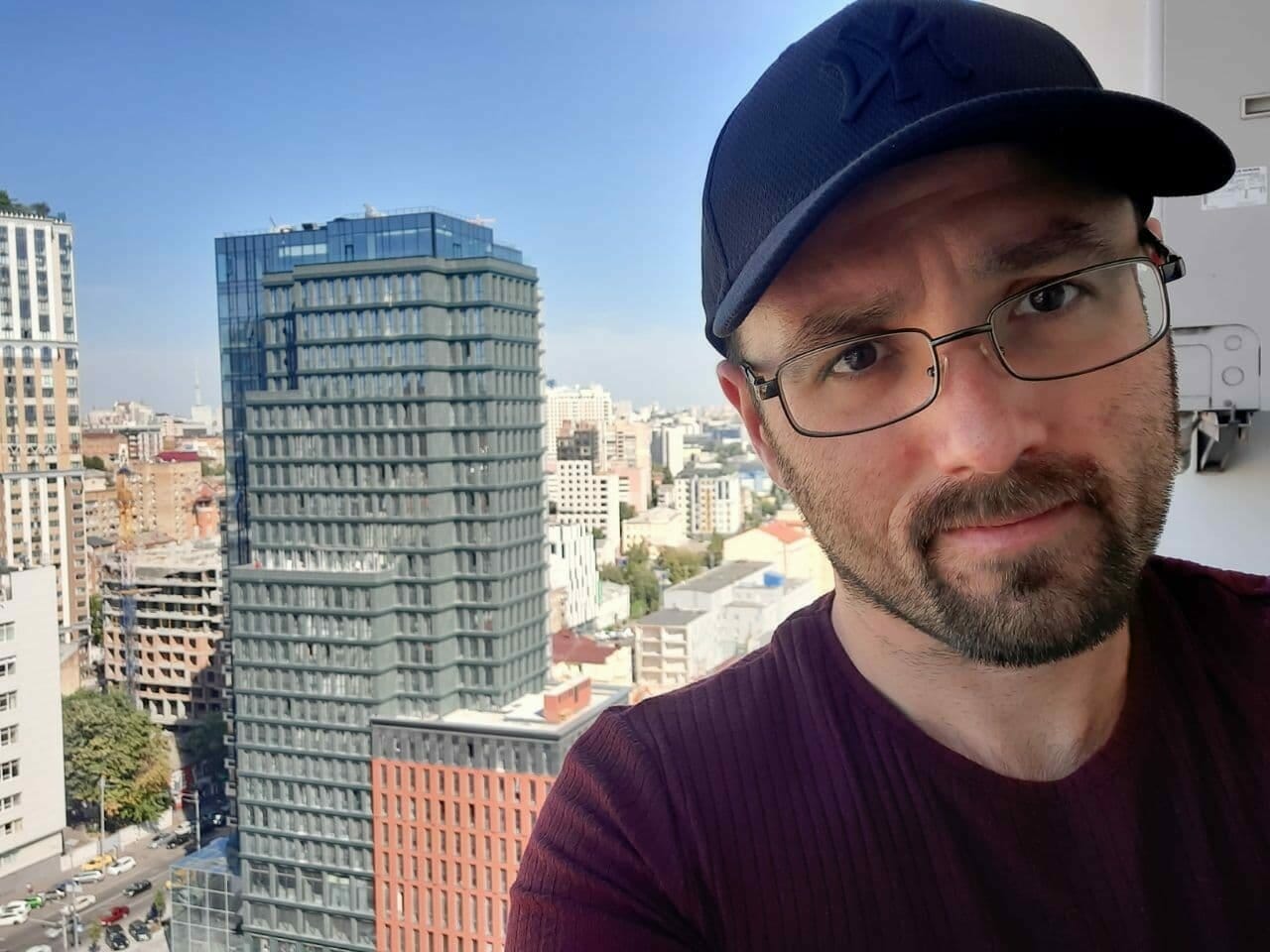 A man wearing a cap and glasses takes a selfie with a cityscape featuring tall buildings and a clear blue sky in the background, capturing the vibrant energy of the place where companies come to hire developers.