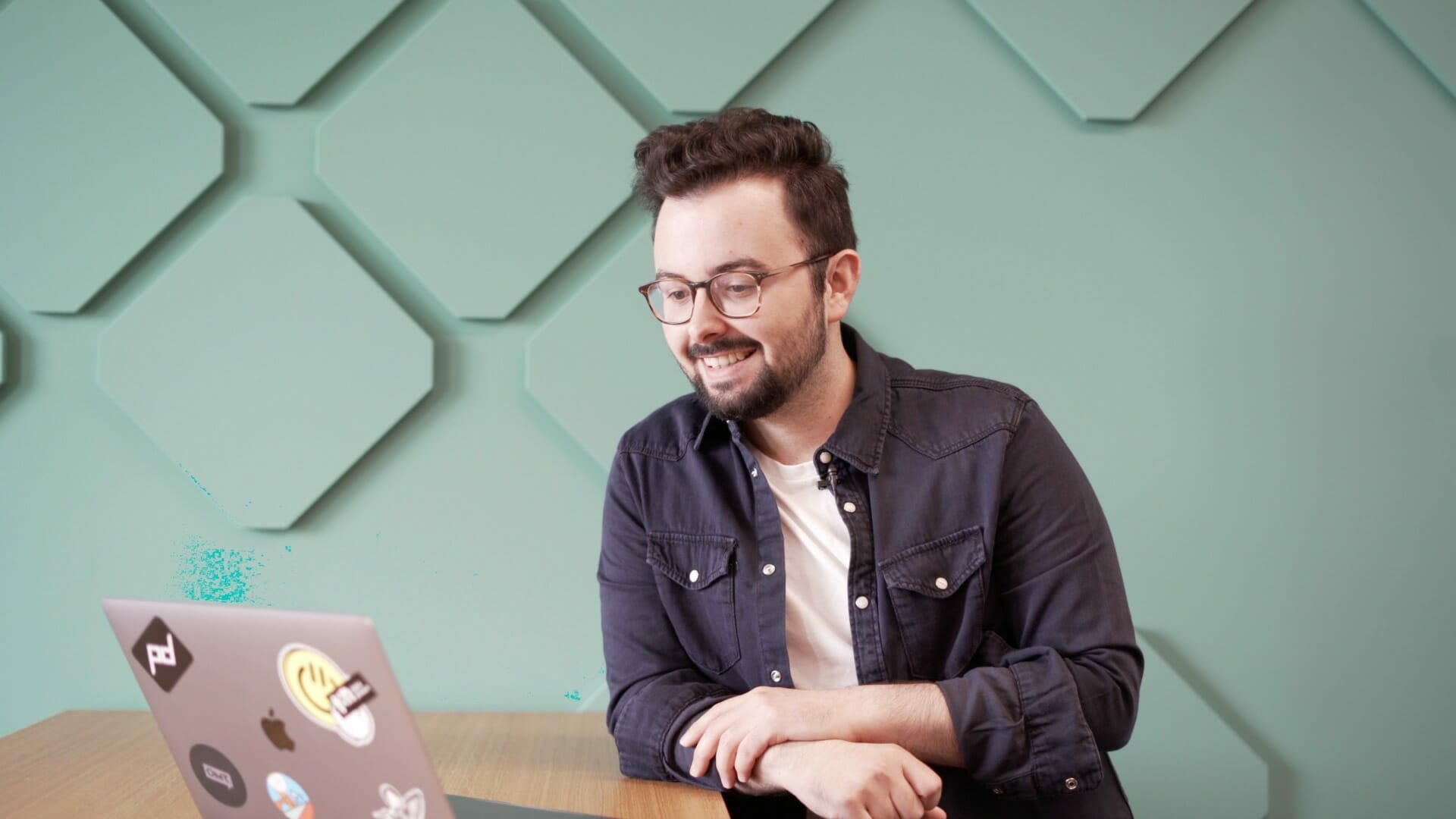 A person sits at a wooden table using a laptop for digital commerce. The background features a teal wall with geometric patterns. The person is wearing glasses, a dark shirt, and has a slight smile.